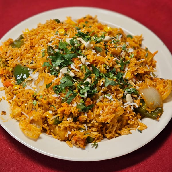 A plate of vegetable biryani with rice, green peppers, onions, cashews, and cilantro on a red tablecloth.