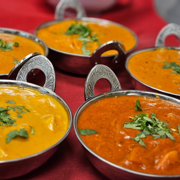 Five bowls of Indian curry, some orange and some red, garnished with cilantro on a red tablecloth.