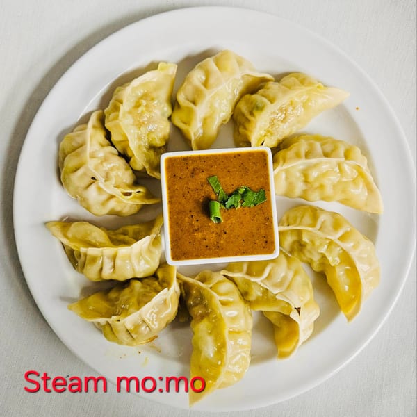 A white plate of steamed momos arranged around a square dish of orange dipping sauce.