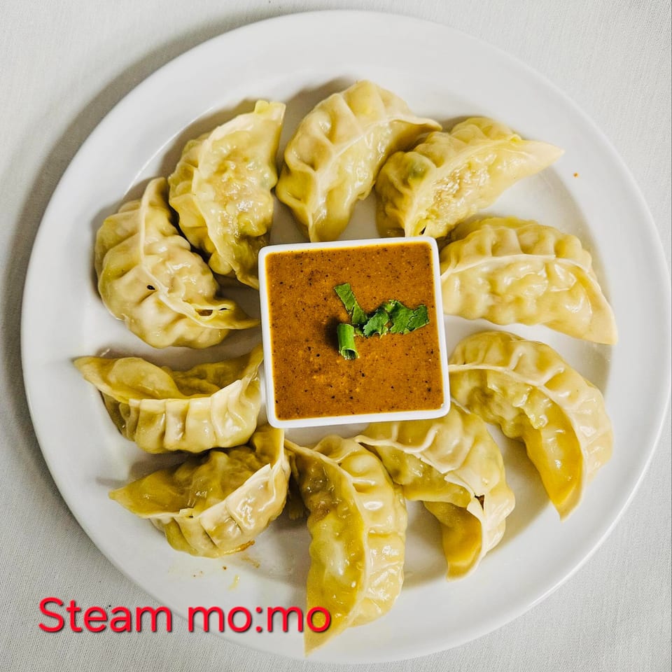 A white plate of steamed momos arranged around a square dish of orange dipping sauce.